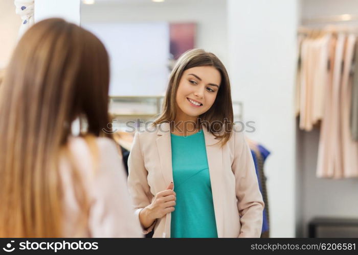 shopping, clothes, fashion, style and people concept - happy woman choosing jacket and posing at mirror in mall or clothing store