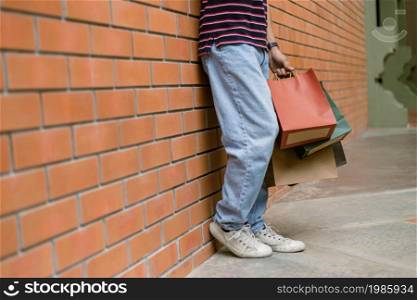 shopping concept A person in light blue jeans and white shoes leaning on the wall of the building while holding a lot of paper shopping bags.