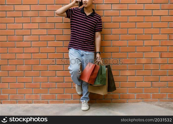 shopping concept The guy in a happy face attempting to make a phone call while slanting himself on the wall and bringing plenty of shopping bags with him.