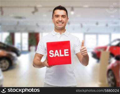 shopping, discount, consumerism, gesture and people concept - smiling man with red sale sigh showing thumbs up over auto show background