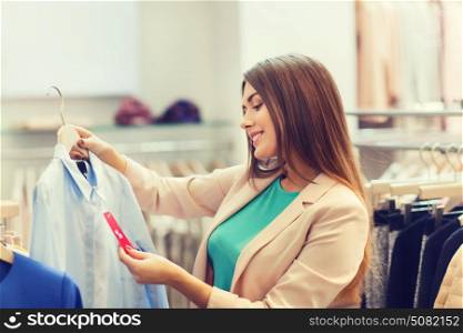 shopping, fashion, clothes, style and people concept - happy young woman looking at shirt price tag label in mall or clothing store. happy young woman choosing clothes in mall