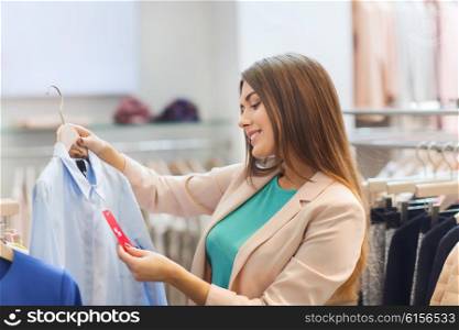 shopping, fashion, clothes, style and people concept - happy young woman looking at shirt price tag label in mall or clothing store