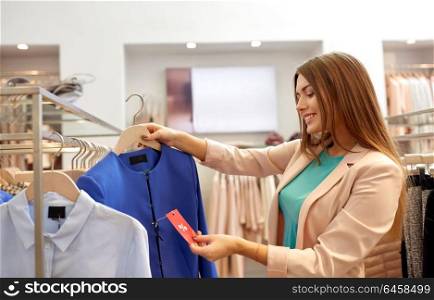 shopping, fashion, sale and people concept - happy young woman choosing clothes in mall or clothing store and looking at discount tag. happy woman choosing clothes at clothing store