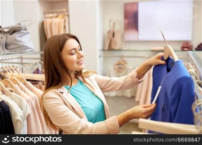 shopping, fashion, sale and people concept - happy young woman choosing clothes in mall or clothing store and looking at price tag. happy woman choosing clothes at clothing store. happy woman choosing clothes at clothing store