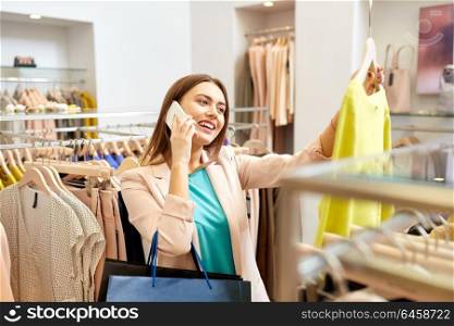 shopping, fashion, sale and people concept - happy young woman choosing dress and calling on smartphone in mall or clothing store. woman calling on smartphone at clothing store