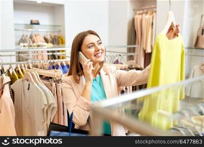 shopping, fashion, sale and people concept - happy young woman choosing dress and calling on smartphone in mall or clothing store. woman calling on smartphone at clothing store. woman calling on smartphone at clothing store