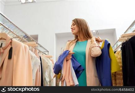 shopping, fashion, sale and people concept - happy young woman with clothes on hangers in mall or clothing store. happy young woman choosing clothes in mall