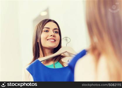 shopping, fashion, style and people concept - happy woman choosing clothes and looking to mirror in mall or clothing store. happy woman with clothes at clothing store mirror