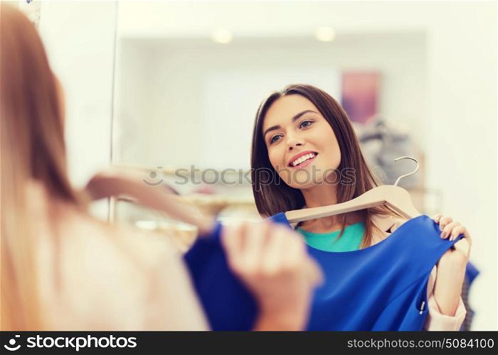 shopping, fashion, style and people concept - happy woman choosing clothes and looking to mirror in mall or clothing store. happy woman with clothes at clothing store mirror. happy woman with clothes at clothing store mirror