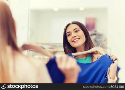 shopping, fashion, style and people concept - happy woman choosing clothes and looking to mirror in mall or clothing store. happy woman with clothes at clothing store mirror. happy woman with clothes at clothing store mirror