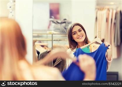 shopping, fashion, style and people concept - happy woman choosing clothes and looking to mirror in mall or clothing store. happy woman with clothes at clothing store mirror