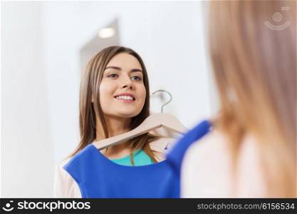 shopping, fashion, style and people concept - happy woman choosing clothes and looking to mirror in mall or clothing store