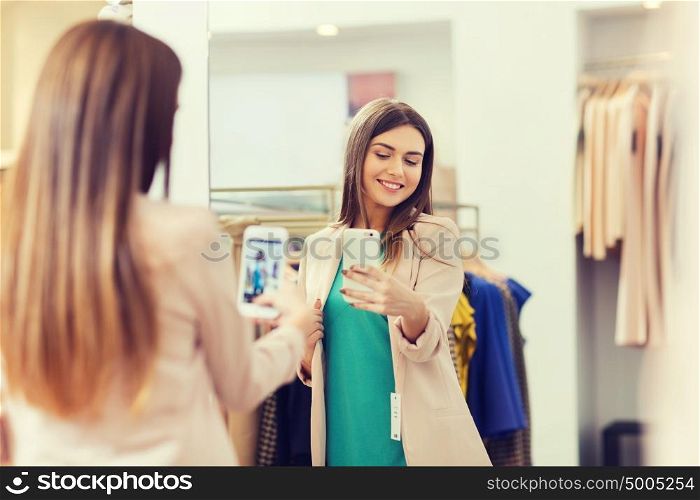 shopping, fashion, style, technology and people concept - happy woman with smartphone taking mirror selfie at clothing store. woman taking mirror selfie by smartphone at store