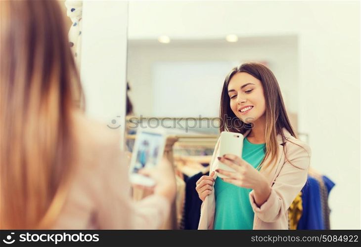 shopping, fashion, style, technology and people concept - happy woman with smartphone taking mirror selfie at clothing store. woman taking mirror selfie by smartphone at store. woman taking mirror selfie by smartphone at store