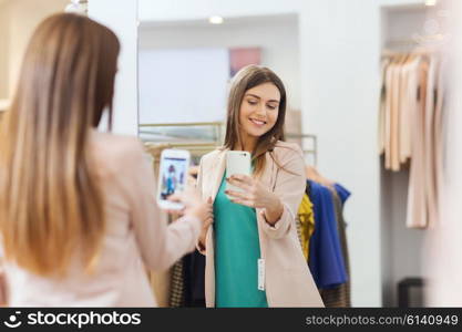 shopping, fashion, style, technology and people concept - happy woman with smartphone taking mirror selfie at clothing store