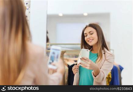 shopping, fashion, style, technology and people concept - happy woman with smartphone taking mirror selfie at clothing store