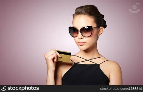 shopping, finances, fashion, people and luxury concept - beautiful young woman in elegant black sunglasses with credit card over violet background