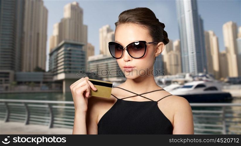 shopping, finances, fashion, people and luxury concept - beautiful young woman in elegant black sunglasses with credit card over dubai city port background