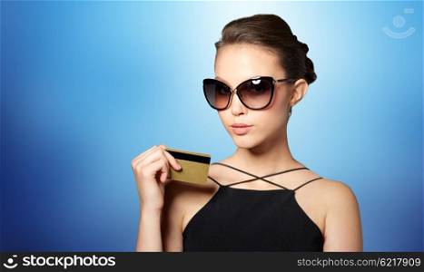 shopping, finances, fashion, people and luxury concept - beautiful young woman in elegant black sunglasses with credit card over blue background