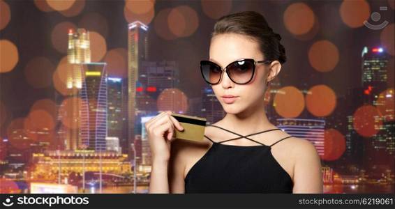 shopping, finances, fashion, people and luxury concept - beautiful young woman in elegant black sunglasses with credit card over night city lights background