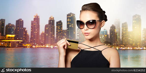 shopping, finances, fashion, people and luxury concept - beautiful young woman in elegant black sunglasses with credit card over night dubai city lights background