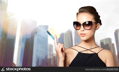 shopping, finances, fashion, people and luxury concept - beautiful young woman in elegant black sunglasses with credit card over city skyscrapers background