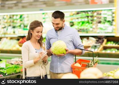 shopping, food, sale, consumerism and people concept - happy young couple with basket and notebook buying cabbage at grocery store or supermarket