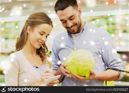 shopping, food, sale, consumerism and people concept - happy young couple with basket and notebook buying cabbage at grocery store or supermarket over snow. couple with notebook and cabbage at grocery store