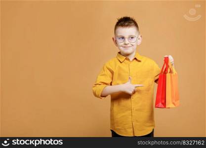 Shopping on black friday. Little boy holding shopping bags on yellow background and showing finger on bag. Shopper with many colored paper bags. Holidays sales and discounts. Cyber Monday. Shopping on black friday. Little boy holding shopping bags on yellow background and showing finger on bag. Shopper with many colored paper bags. Holidays sales and discounts. Cyber Monday.