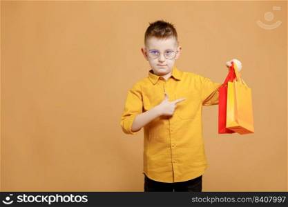 Shopping on black friday. Little boy holding shopping bags on yellow background and showing finger on bag. Shopper with many colored paper bags. Holidays sales and discounts. Cyber Monday. Shopping on black friday. Little boy holding shopping bags on yellow background and showing finger on bag. Shopper with many colored paper bags. Holidays sales and discounts. Cyber Monday.