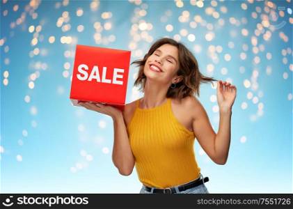 shopping, outlet and people concept - happy smiling young woman in mustard yellow top and jeans with sale sign posing over holidays lights on blue background. happy woman posing with sale sign over lights