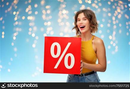 shopping, sale and discount concept - happy smiling young woman in mustard yellow top and jeans with percent sign over holidays lights on blue background. smiling young woman with sale sign over lights