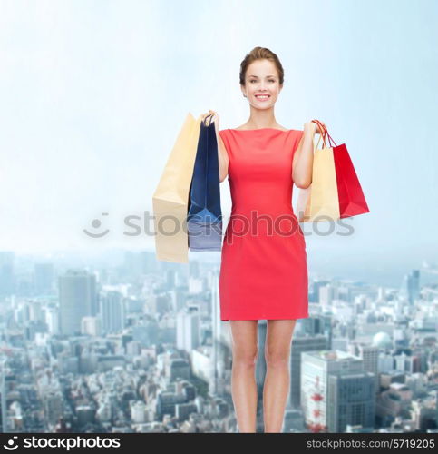 shopping, sale and holidays concept - smiling elegant woman in red dress with shopping bags over city background