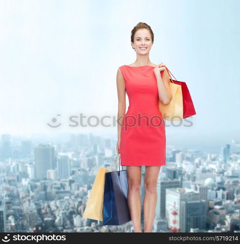 shopping, sale and holidays concept - smiling elegant woman in red dress with shopping bags over city background