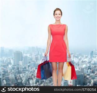 shopping, sale and holidays concept - smiling elegant woman in red dress with shopping bags over city background