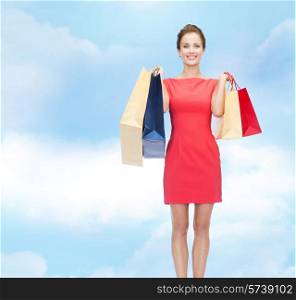 shopping, sale and holidays concept - smiling elegant woman in red dress with shopping bags over blue cloudy sky background
