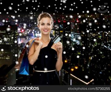 shopping, sale, banking, money and holidays concept - smiling woman in dress with shopping bags and credit card over snowy night city background