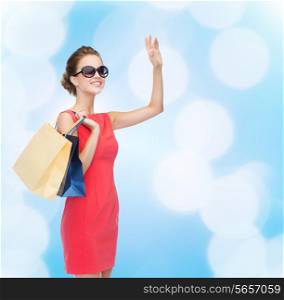 shopping, sale, christmas and holiday concept - smiling elegant woman in red dress and sunglasses with shopping bags waving hand