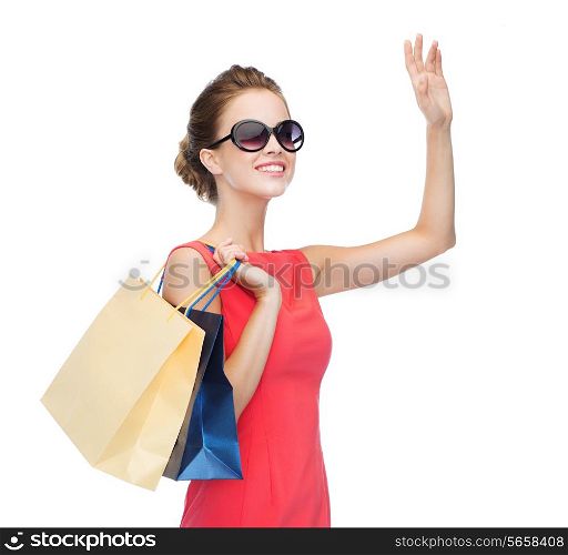 shopping, sale, christmas and holiday concept - smiling elegant woman in red dress and sunglasses with shopping bags waving hand