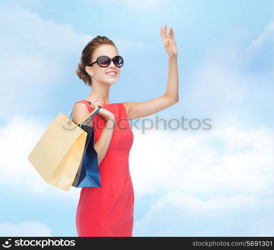 shopping, sale, christmas and holiday concept - smiling elegant woman in red dress and sunglasses with shopping bags waving hand