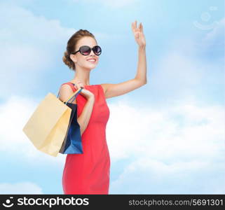 shopping, sale, christmas and holiday concept - smiling elegant woman in red dress and sunglasses with shopping bags waving hand