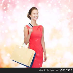 shopping, sale, christmas and holiday concept - smiling elegant woman in red dress with shopping bags and plastic card over pink snowy background