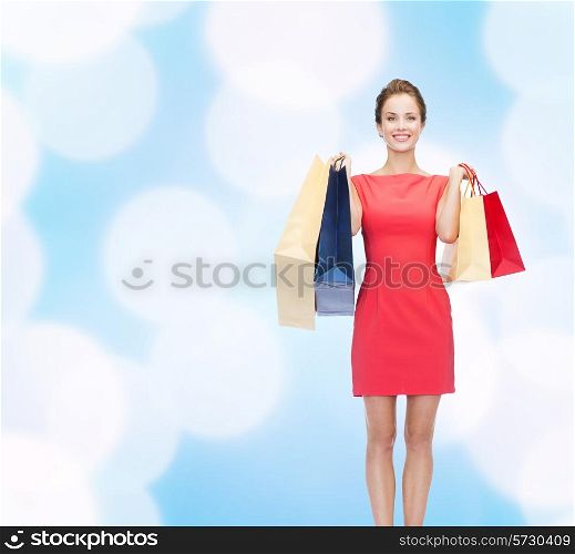 shopping, sale, christmas and holidays concept - smiling elegant woman in red dress with shopping bags over blue lights background