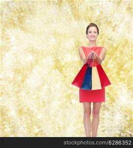 shopping, sale, christmas and holidays concept - smiling elegant woman in red dress with shopping bags over yellow lights background