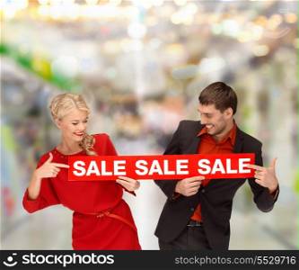 shopping, sale, christmas, couple and mall concept - smiling woman and man pointing finger to red sale sign at shopping mall