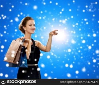 shopping, sale, christmas, people and holidays concept - smiling woman in evening dress with shopping bags pointing finger over blue snowy background