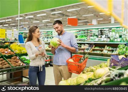 shopping, sale, consumerism and people concept - happy young couple with food basket and notebook at grocery store or supermarket