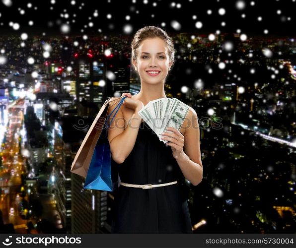 shopping, sale, gifts, money and holidays concept - smiling woman in dress with shopping bags and money over snowy night city background