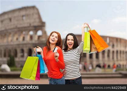 shopping, sale, tourism and people concept - two smiling teenage girls with shopping bags and credit card over coliseum background