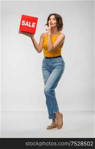 shopping, secret and people concept - happy smiling young woman in mustard yellow top and jeans with sale sign posing and making hush gesture over grey background. happy smiling young woman posing with sale sign